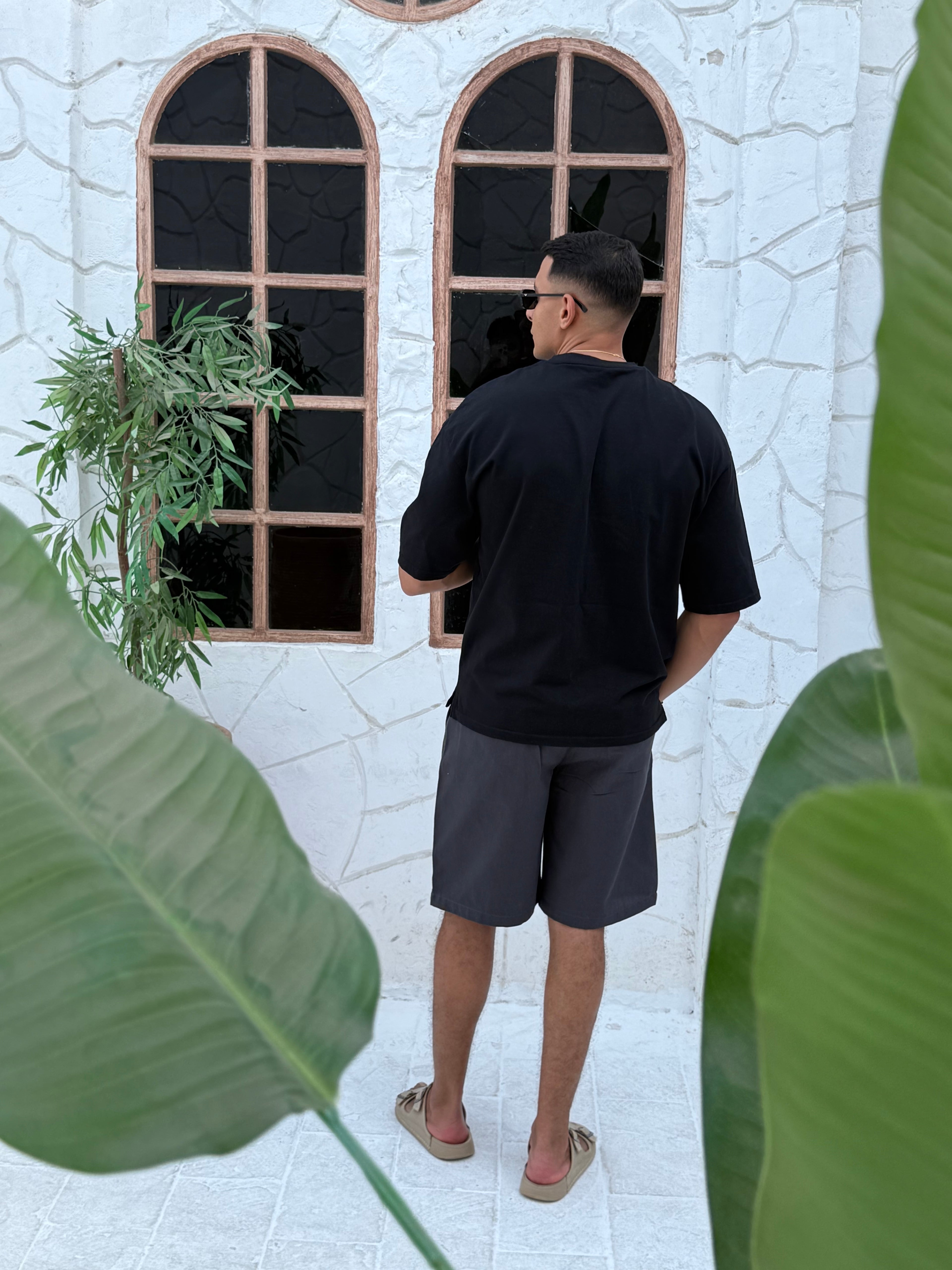 fame clothes A man wearing a black cotton t-shirt, white shorts, and beige sandals stands in front of a white textured stucco building with arched windows. From fame clothes 