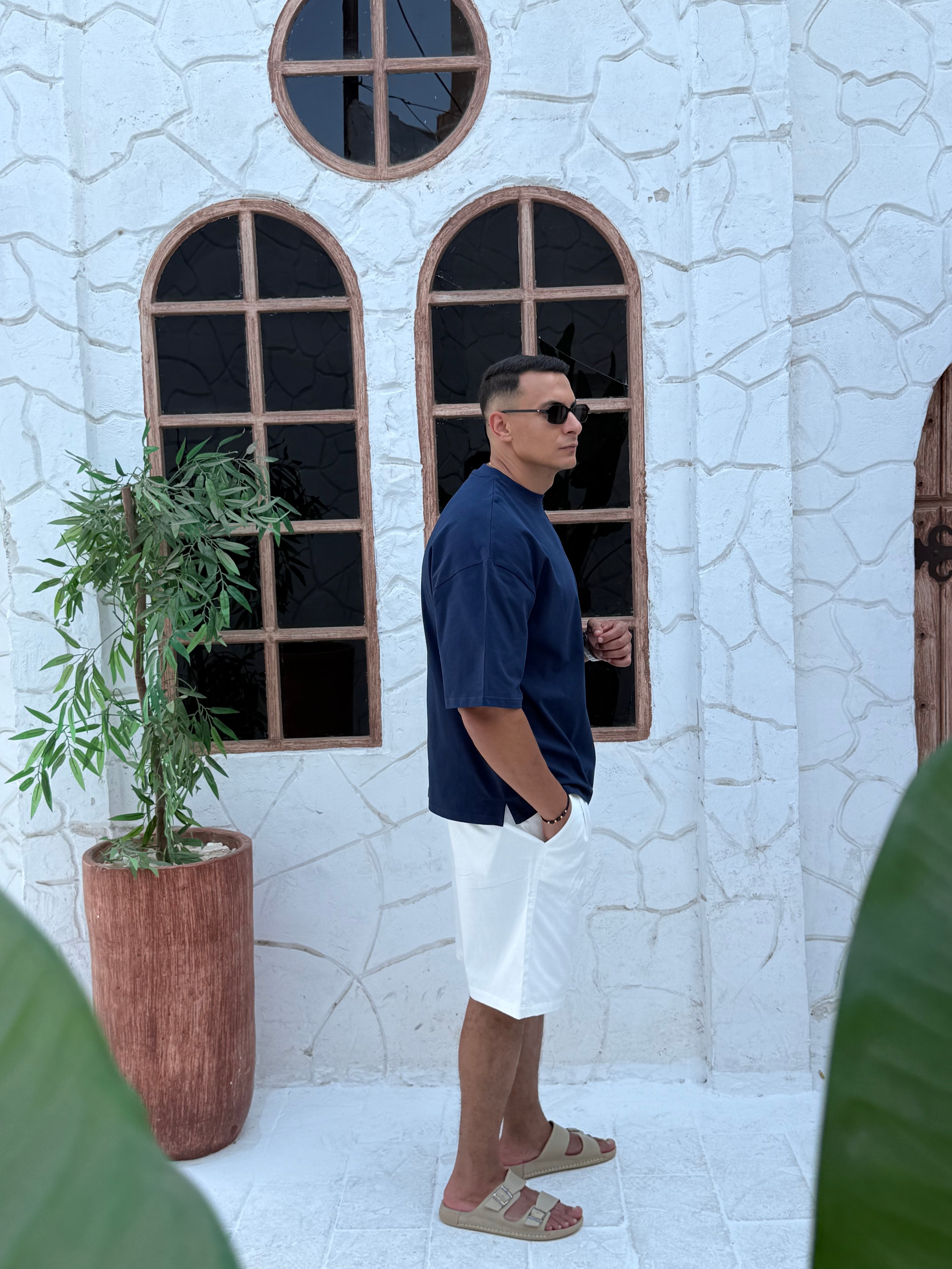 man, wear t-shirt white cotton, fame clothes A man wearing a black cotton t-shirt, white shorts, and beige sandals stands in front of a white textured stucco building with arched windows. From fame clothes 