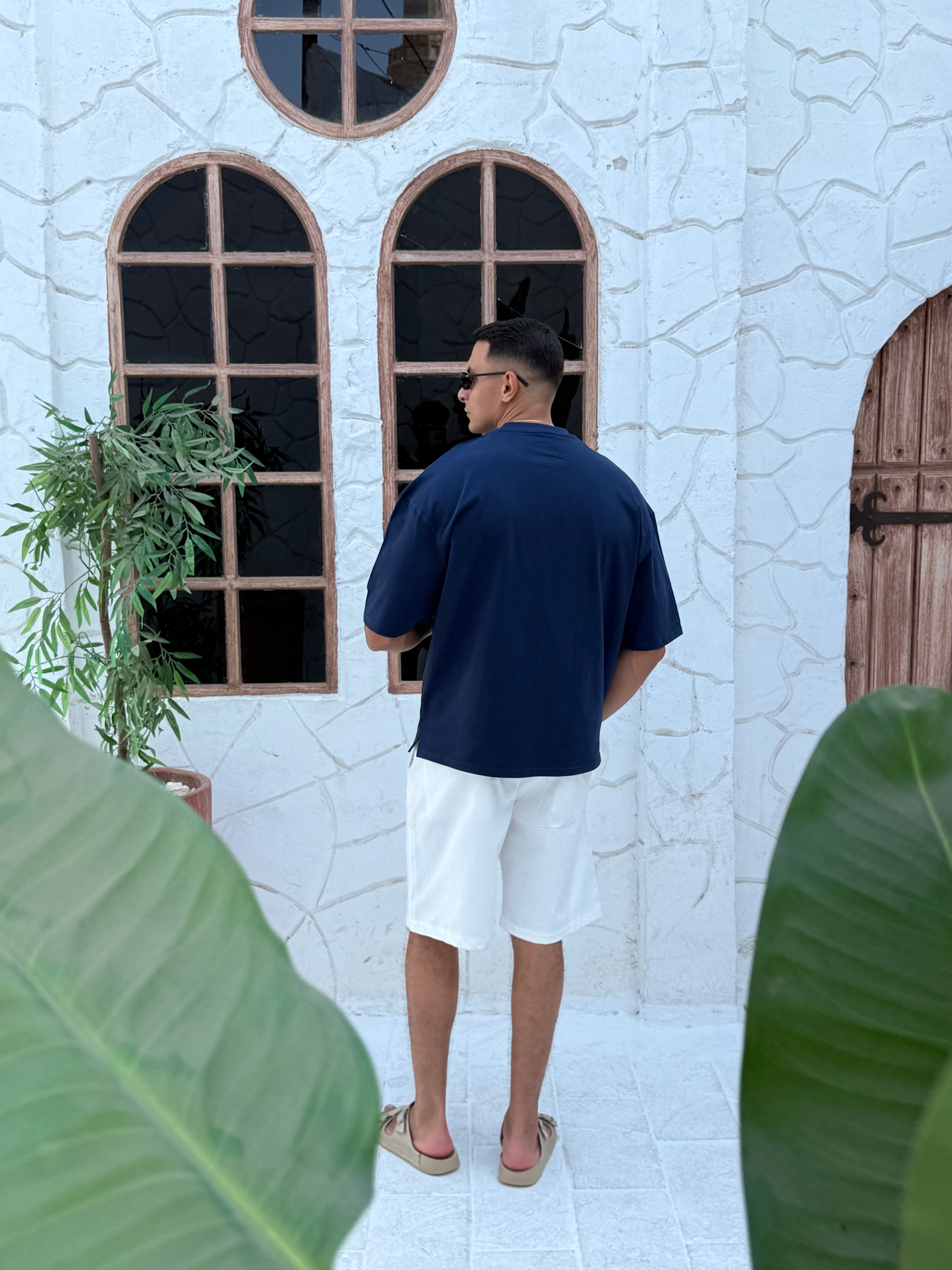 man, wear t-shirt white cotton, fame clothes A man wearing a black cotton t-shirt, white shorts, and beige sandals stands in front of a white textured stucco building with arched windows. From fame clothes 