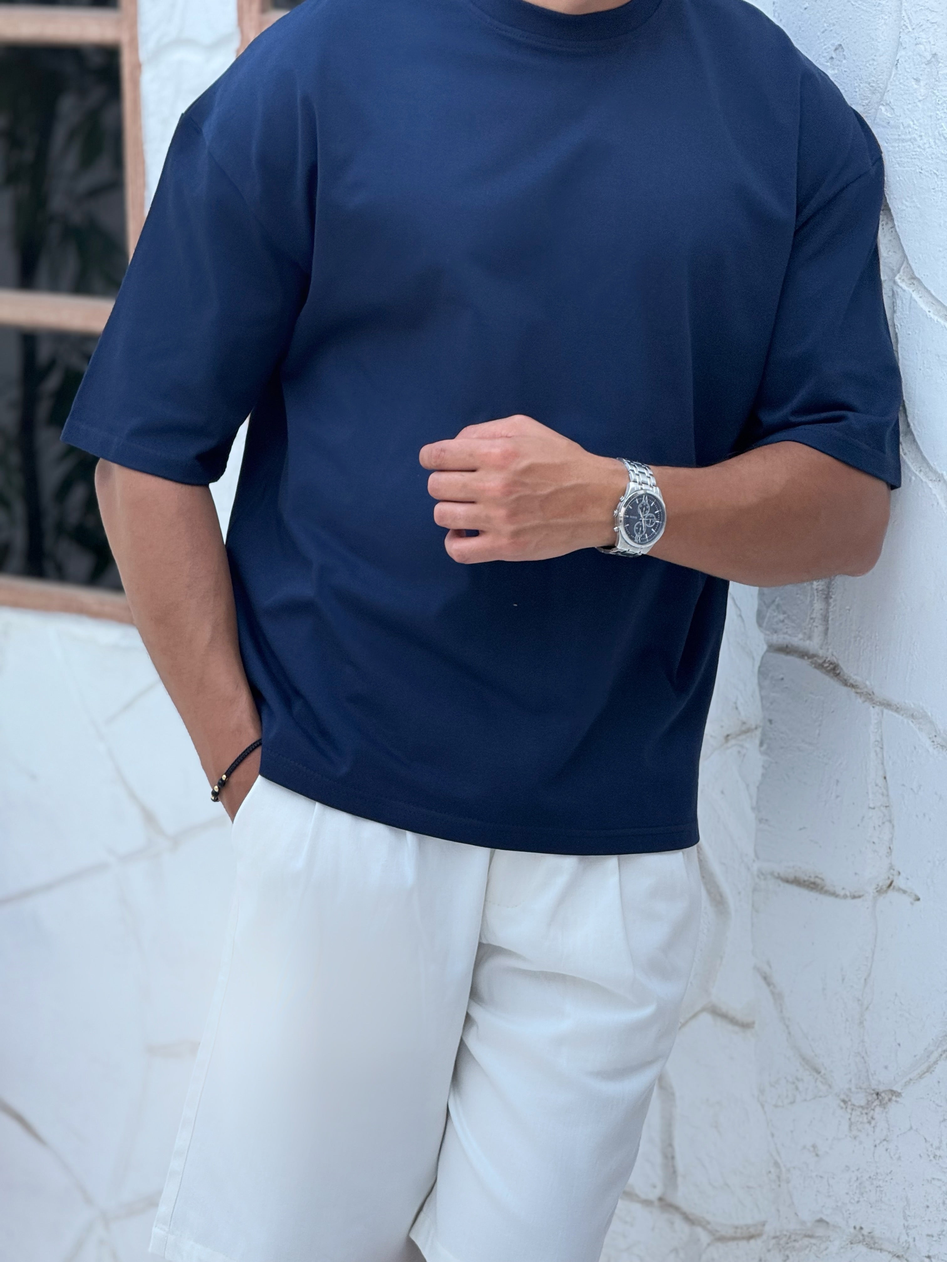man, wear t-shirt white cotton, fame clothes A man wearing a black cotton t-shirt, white shorts, and beige sandals stands in front of a white textured stucco building with arched windows. From fame clothes 