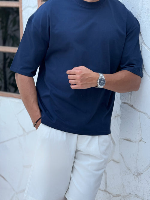 man, wear t-shirt white cotton, fame clothes A man wearing a black cotton t-shirt, white shorts, and beige sandals stands in front of a white textured stucco building with arched windows. From fame clothes 