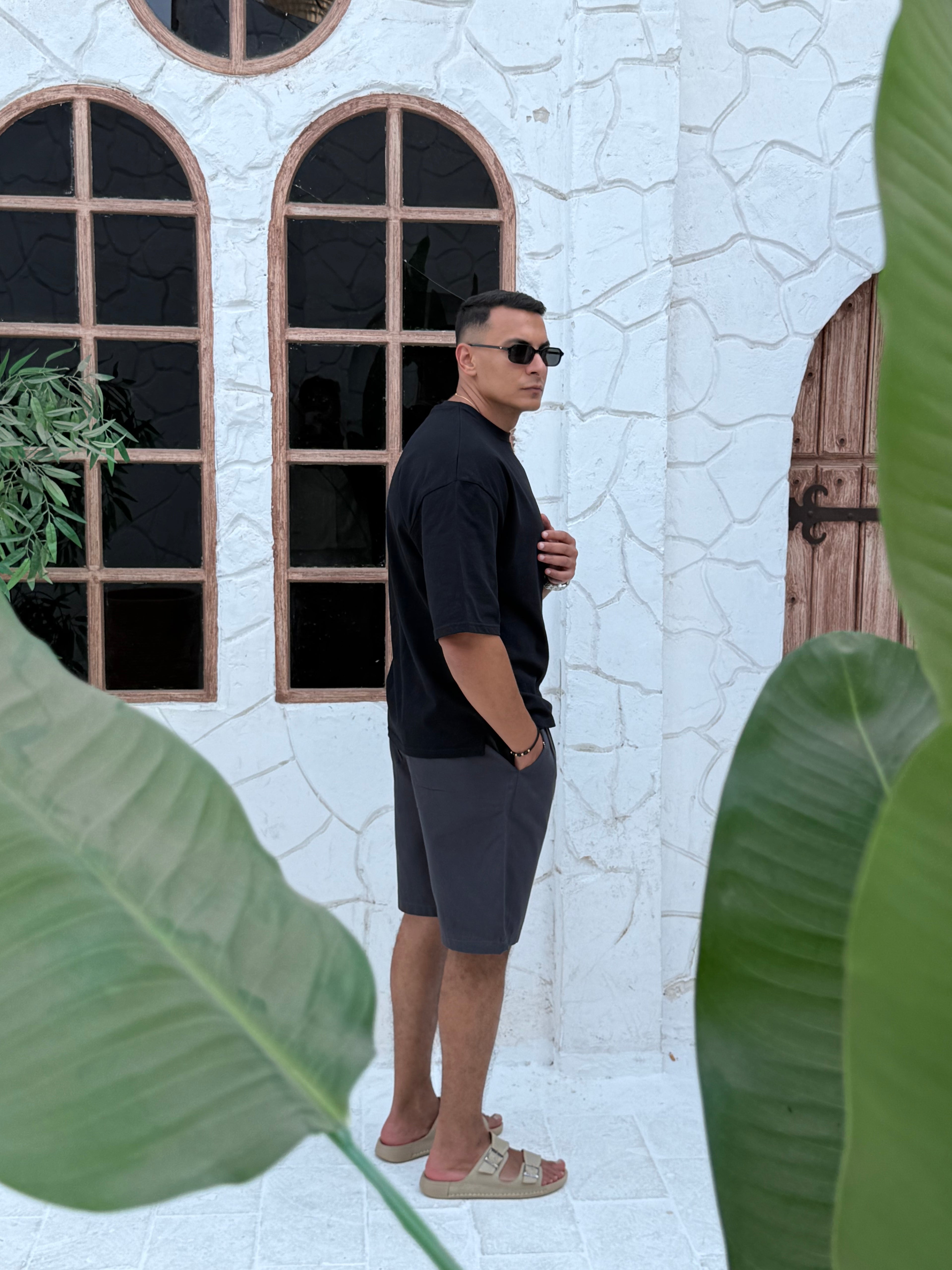 man, wear t-shirt black cotton, fame clothes A man wearing a black cotton t-shirt, white shorts, and beige sandals stands in front of a white textured stucco building with arched windows. From fame clothes 