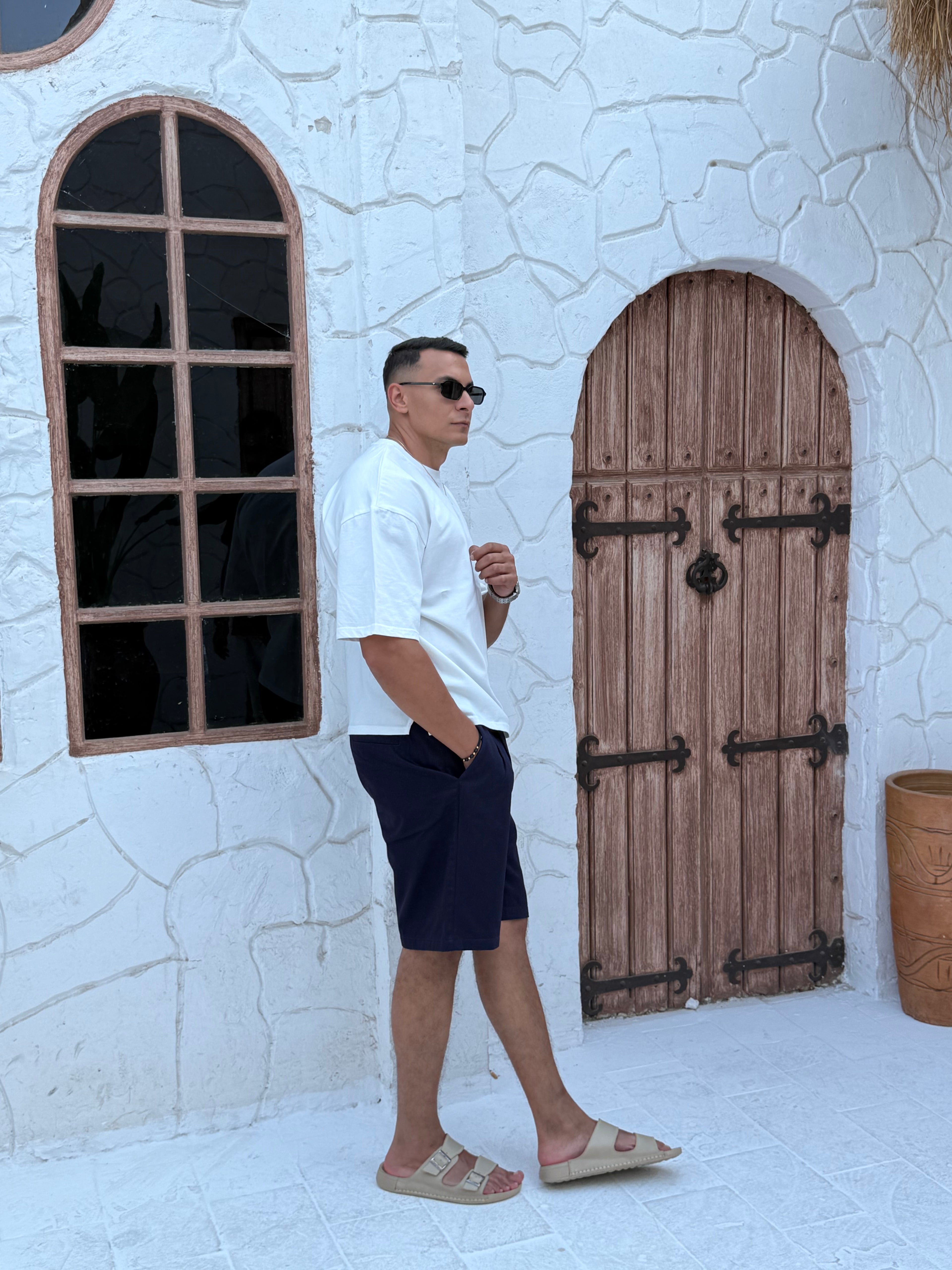 fame clothes A man wearing a navy-blue cotton t-shirt, white shorts, and beige sandals stands in front of a white textured stucco building with arched windows.from fame clothes 