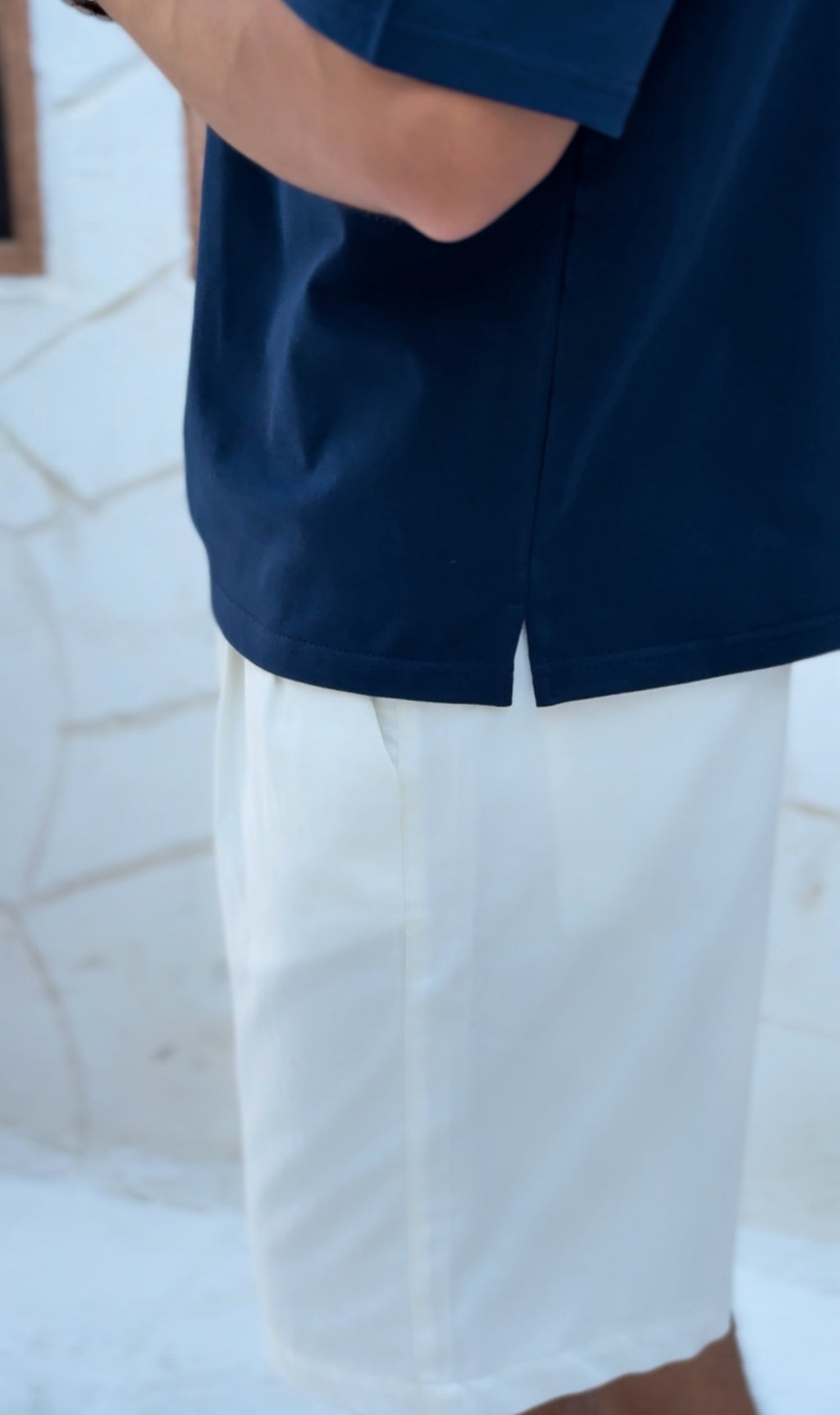 fame clothes A man wearing a black cotton t-shirt, white shorts, and beige sandals stands in front of a white textured stucco building with arched windows. From fame clothes 