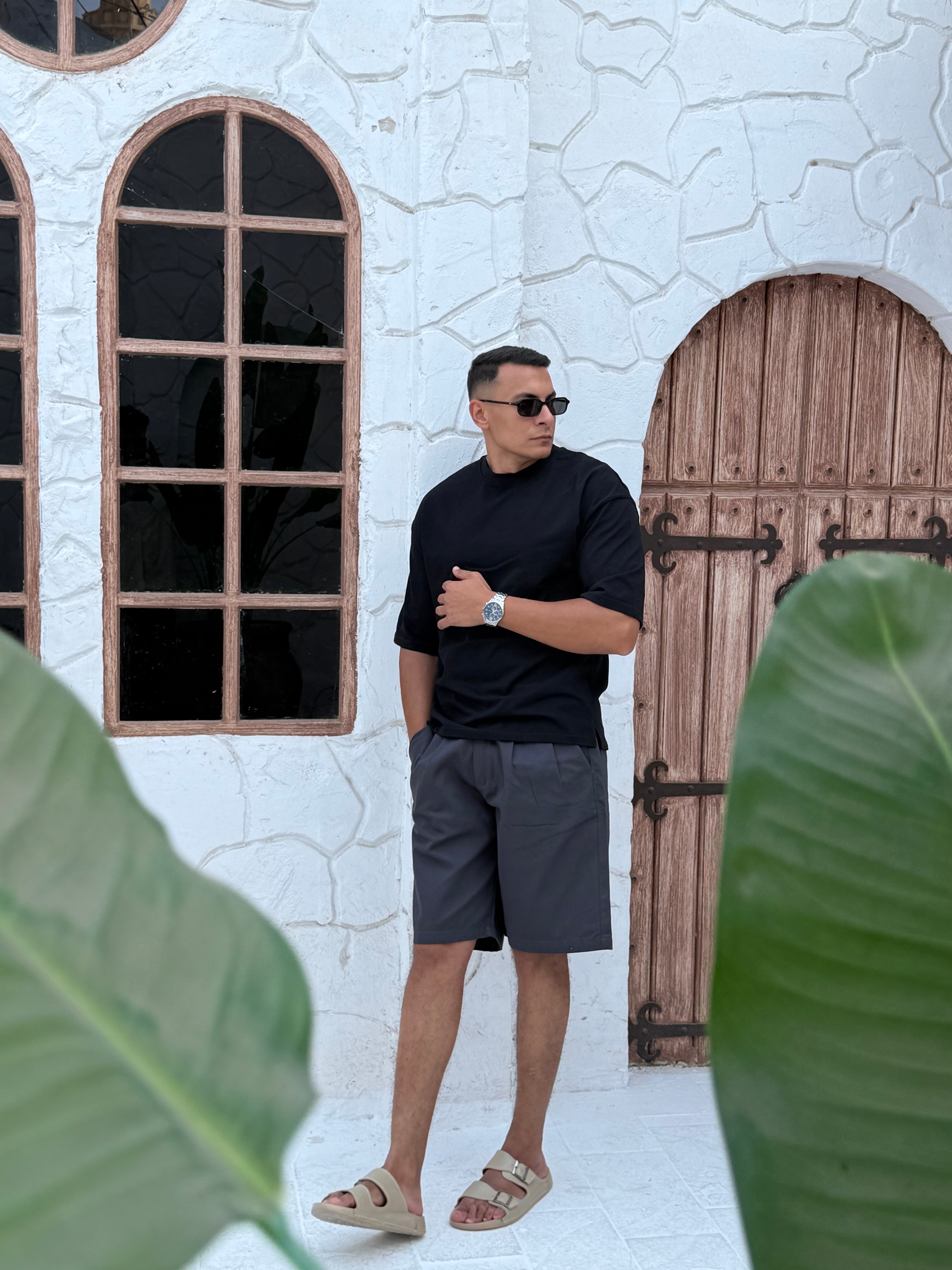 fame clothes A man wearing a black cotton t-shirt, white shorts, and beige sandals stands in front of a white textured stucco building with arched windows. From fame clothes 