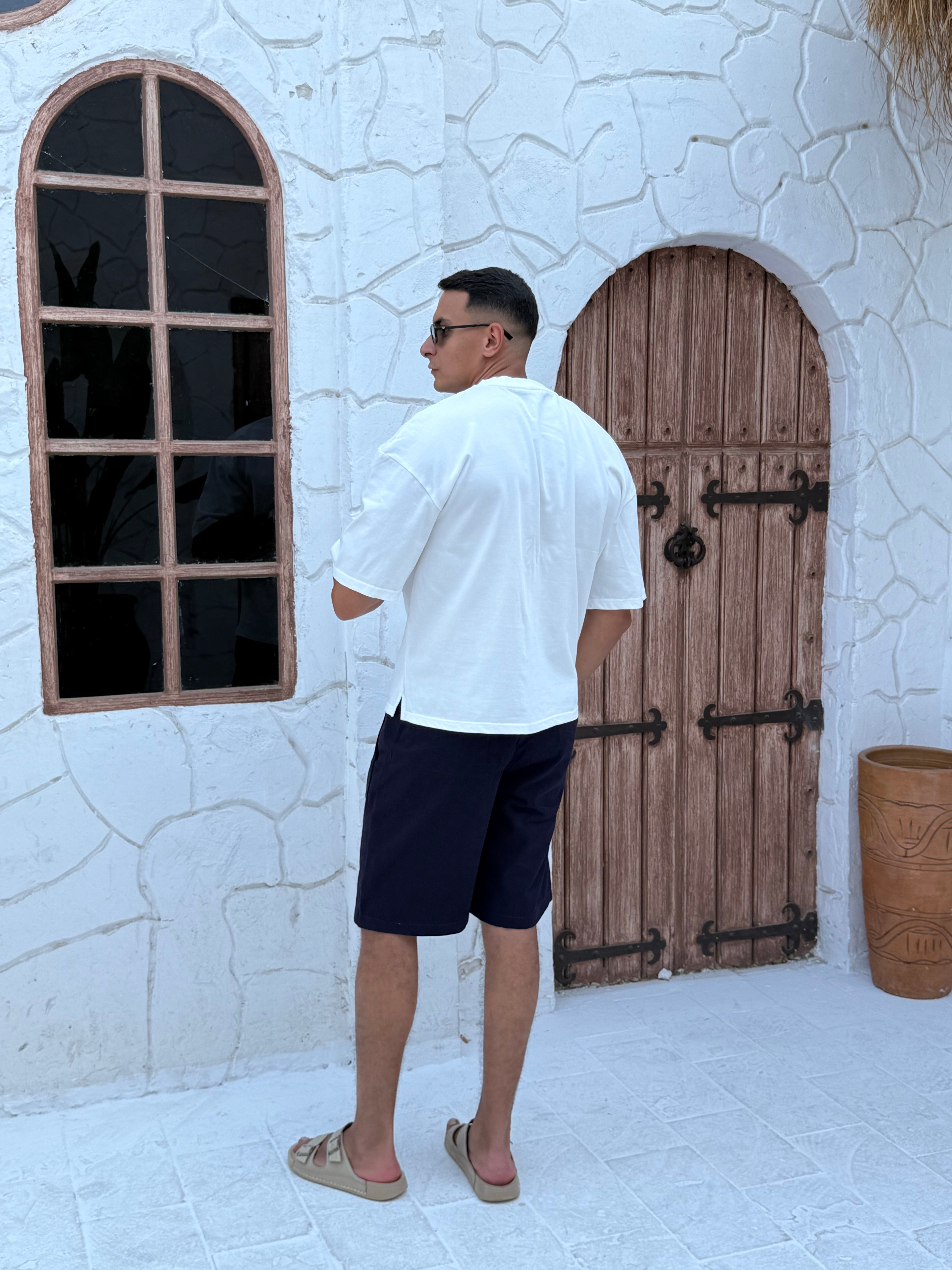 man, wear t-shirt white cotton, fame clothes A man wearing a black cotton t-shirt, white shorts, and beige sandals stands in front of a white textured stucco building with arched windows. From fame clothes 
