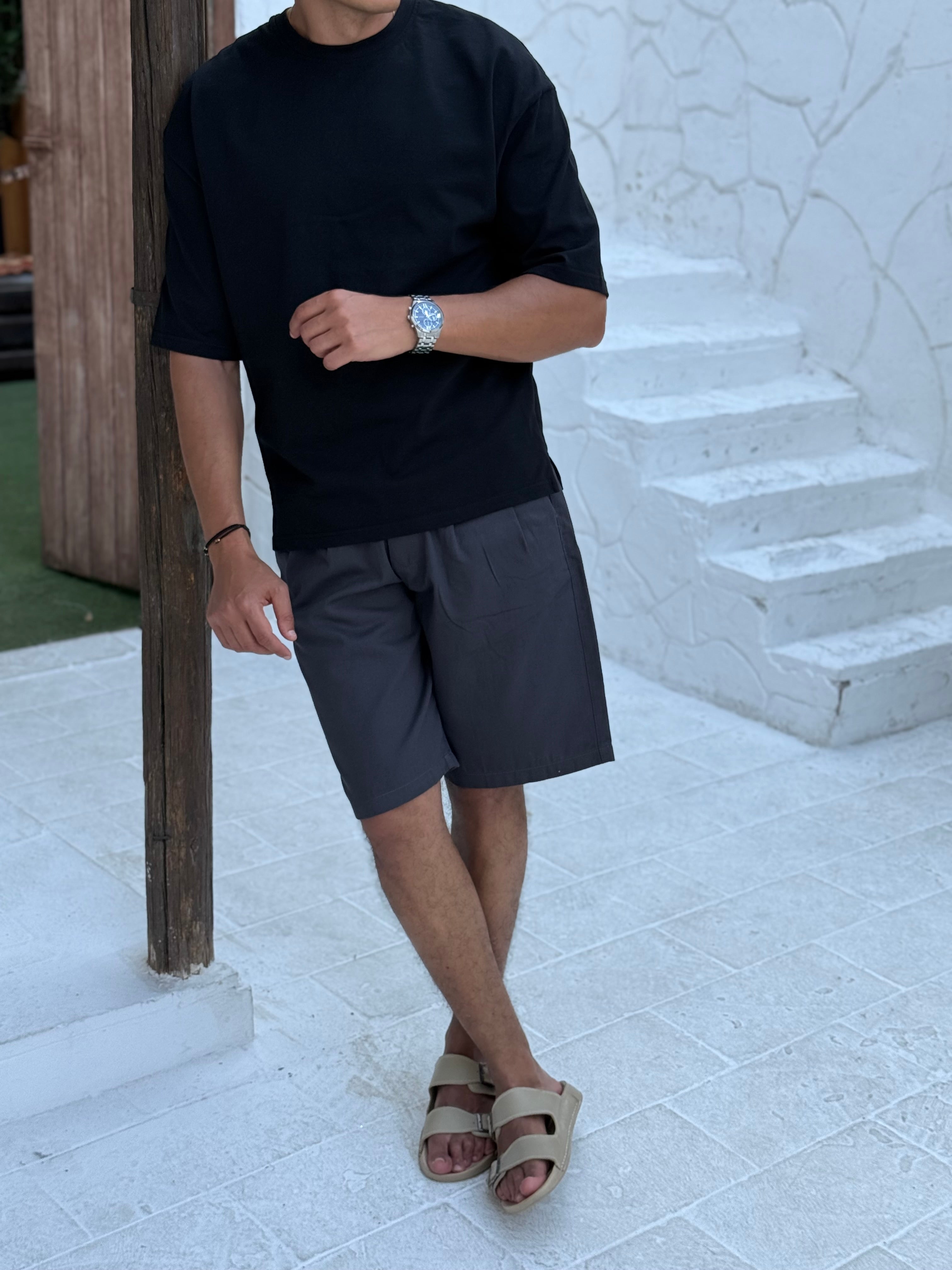 fame clothes A man wearing a black cotton t-shirt, white shorts, and beige sandals stands in front of a white textured stucco building with arched windows. From fame clothes 