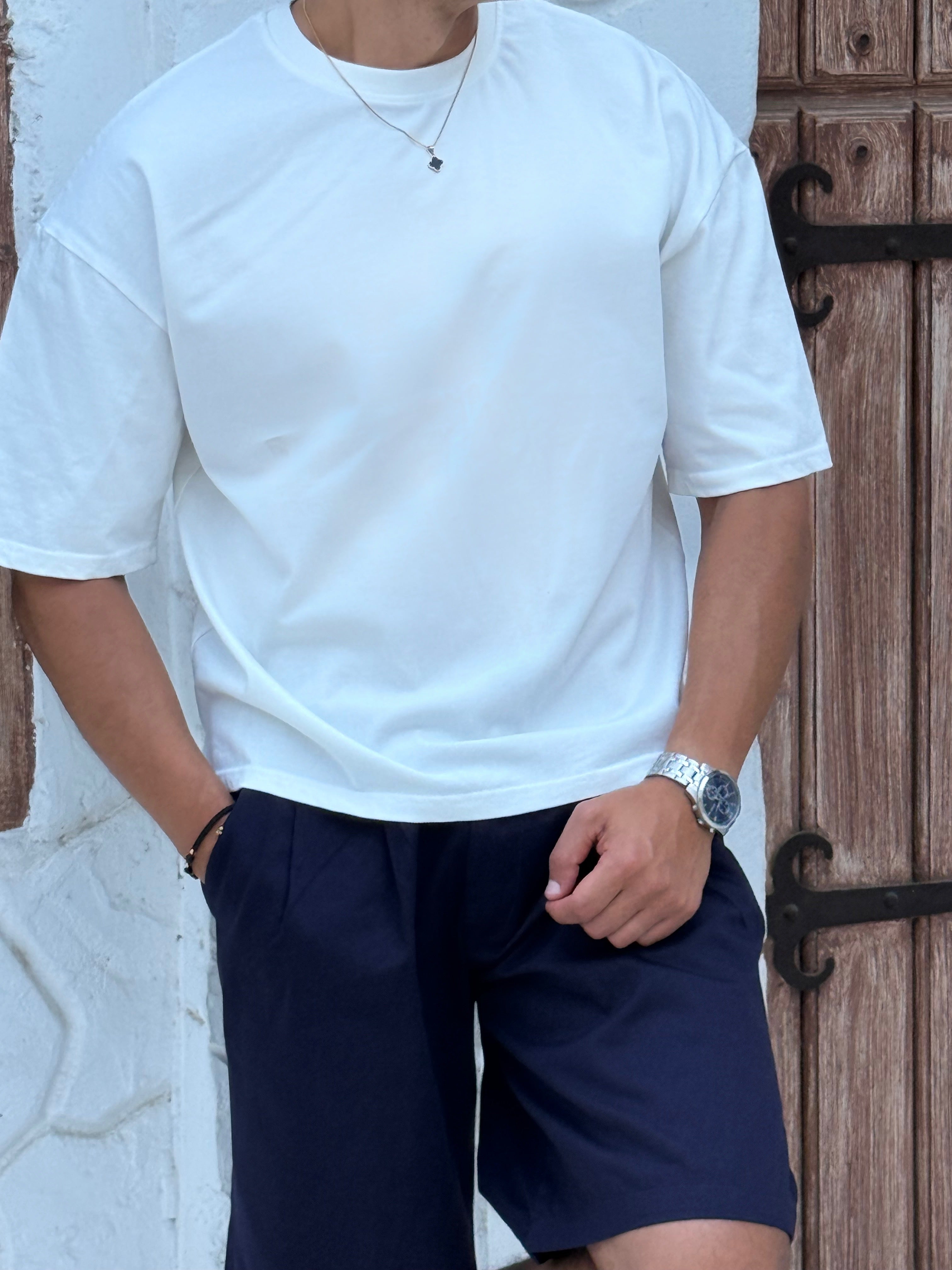 fame clothes A man wearing a black cotton t-shirt, white shorts, and beige sandals stands in front of a white textured stucco building with arched windows. From fame clothes 
